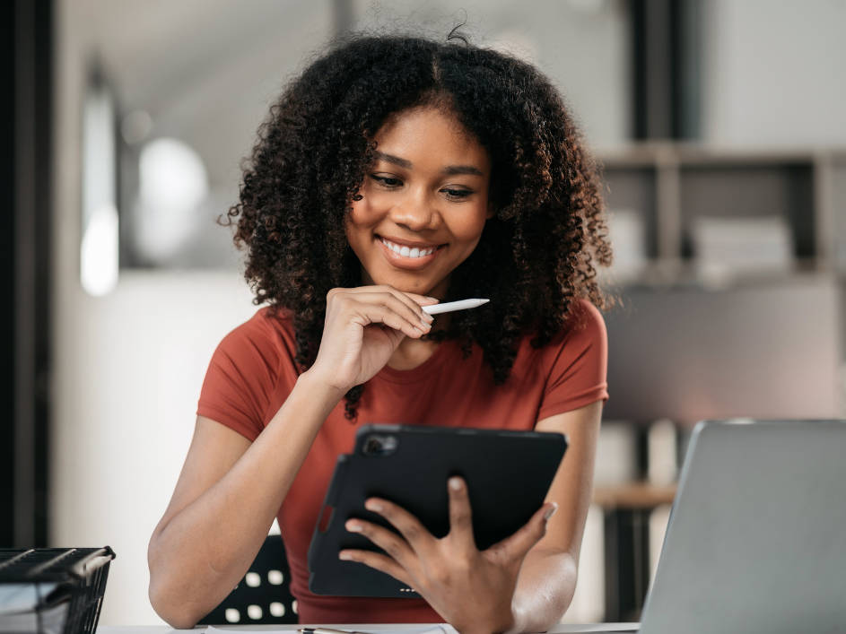 A smiling woman with curly hair holds a stylus and looks at a tablet screen.