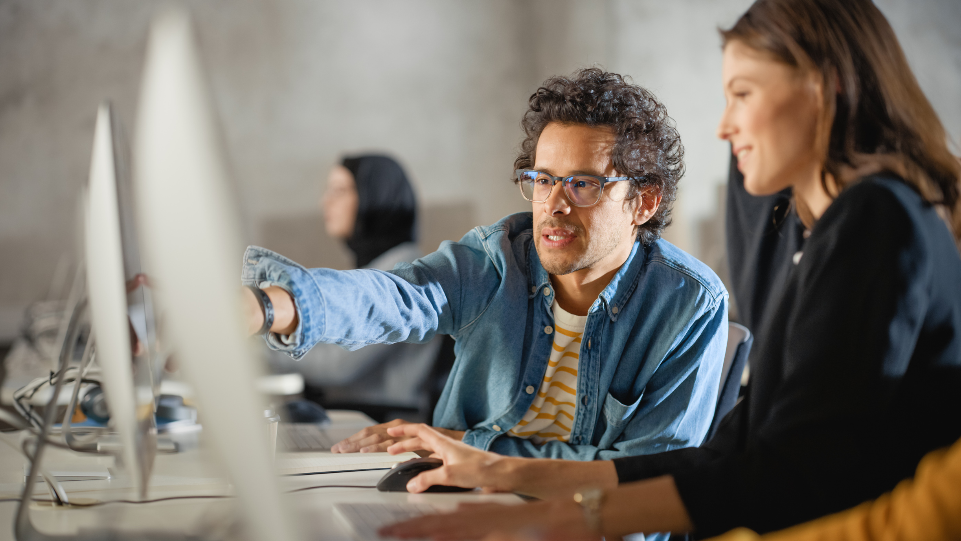 A male adult student in glasses points at a computer monitor while a female student looks on.