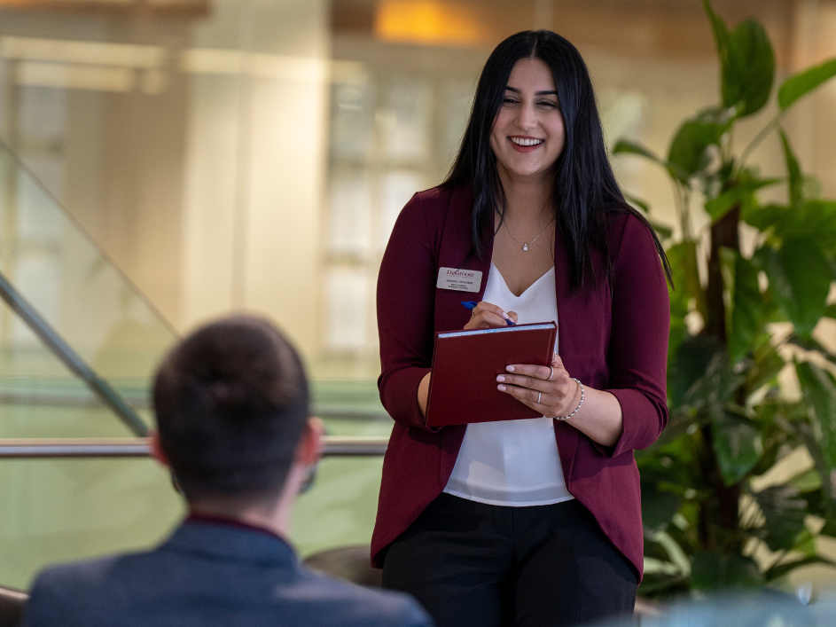 A smiling woman with dark hair and a maroon blazer takes notes while looking at a student.