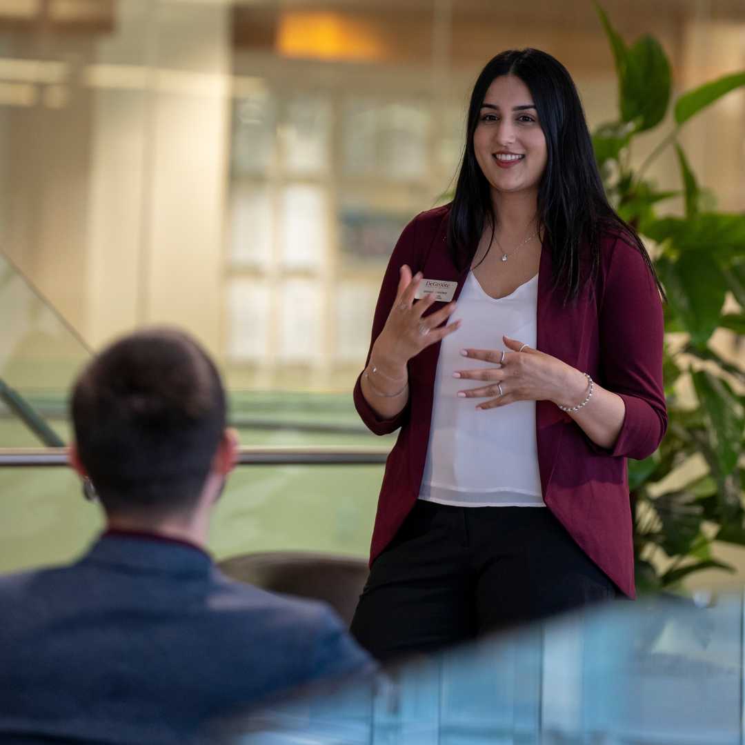 A smiling woman with long dark hair gestures while speaking to a seated man.
