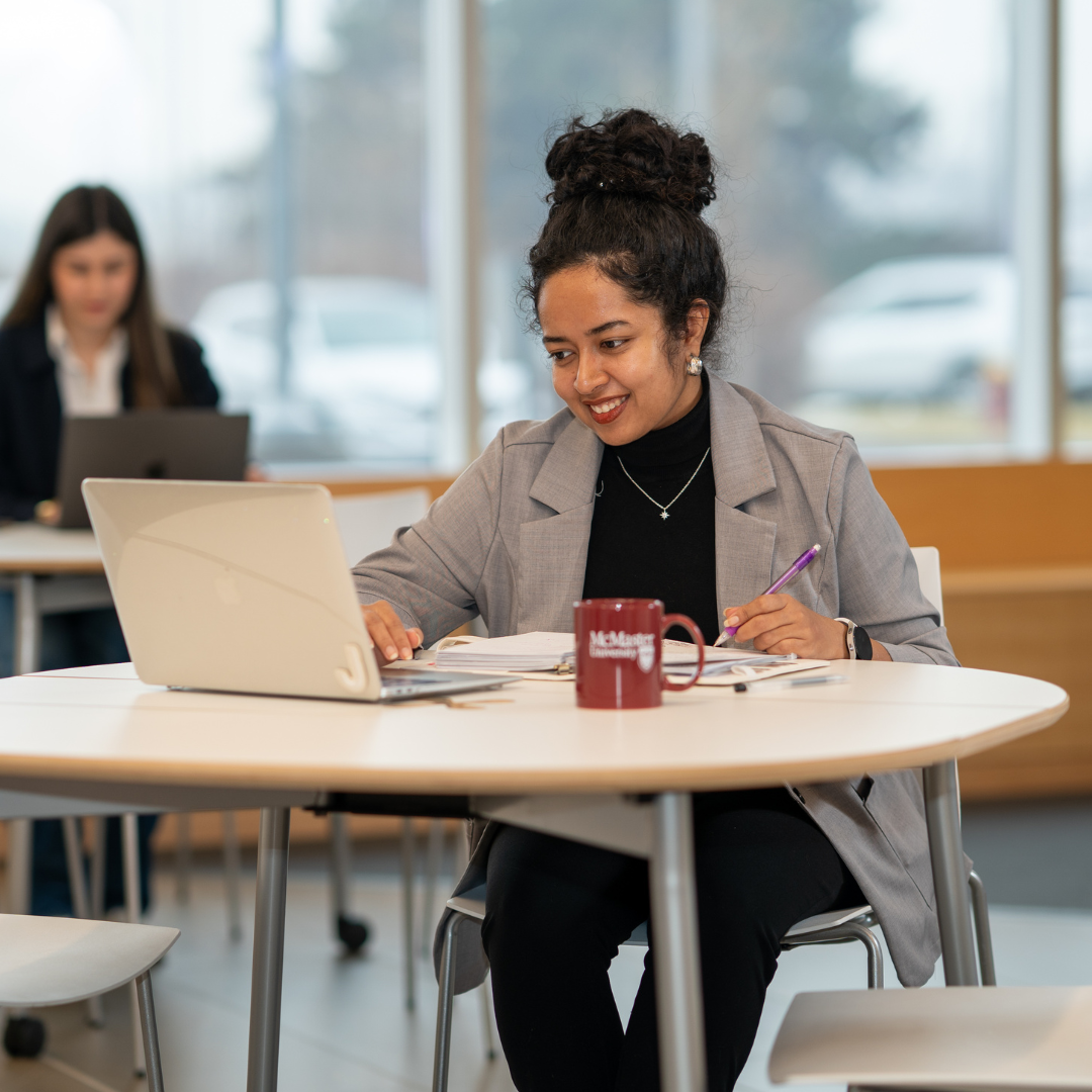 A smiling female DeGroote student with her hair in a bun works on a laptop at a table.