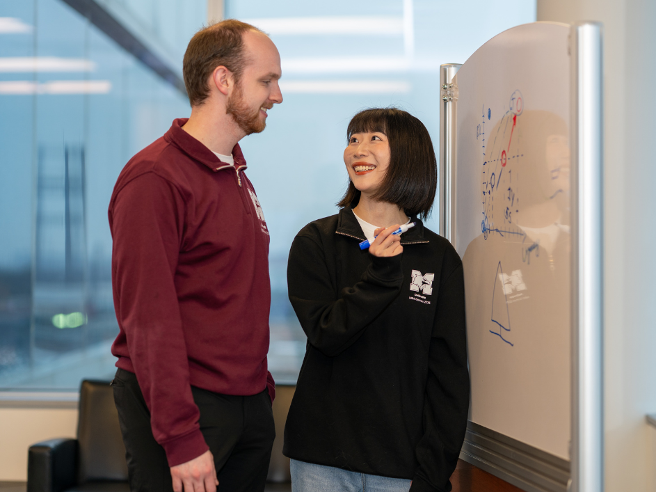 A female student holding up a marker smiles at a male student next to a whiteboard with diagrams.