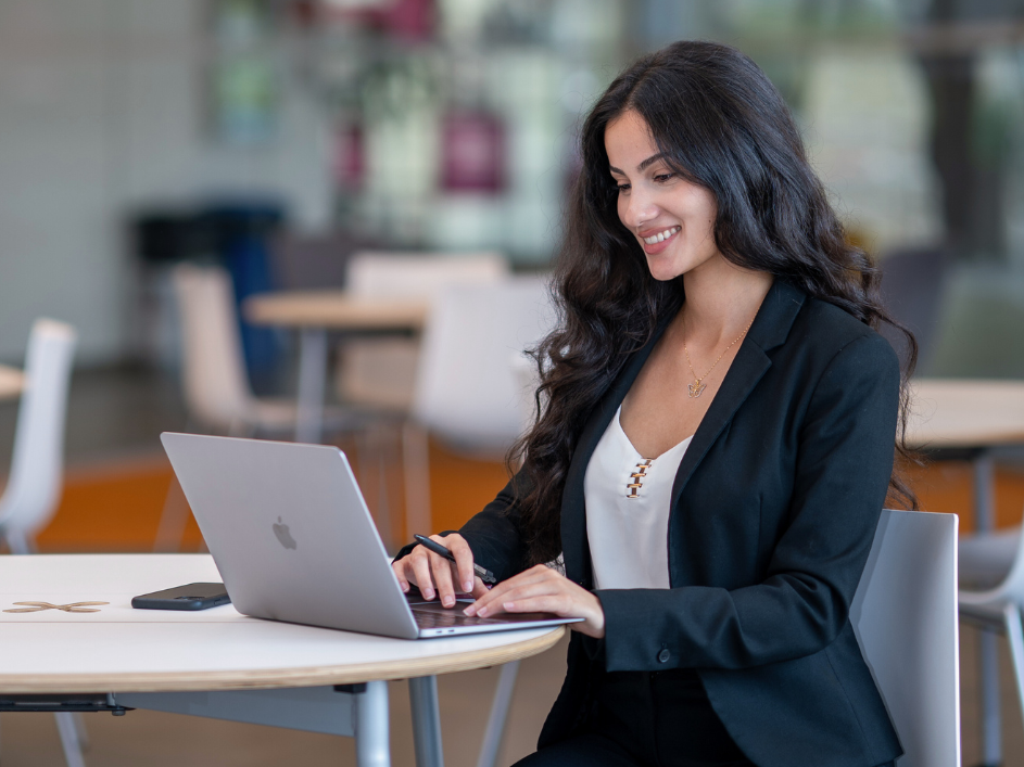 A smiling woman with long dark hair sits at a table, looking at and typing on a laptop.
