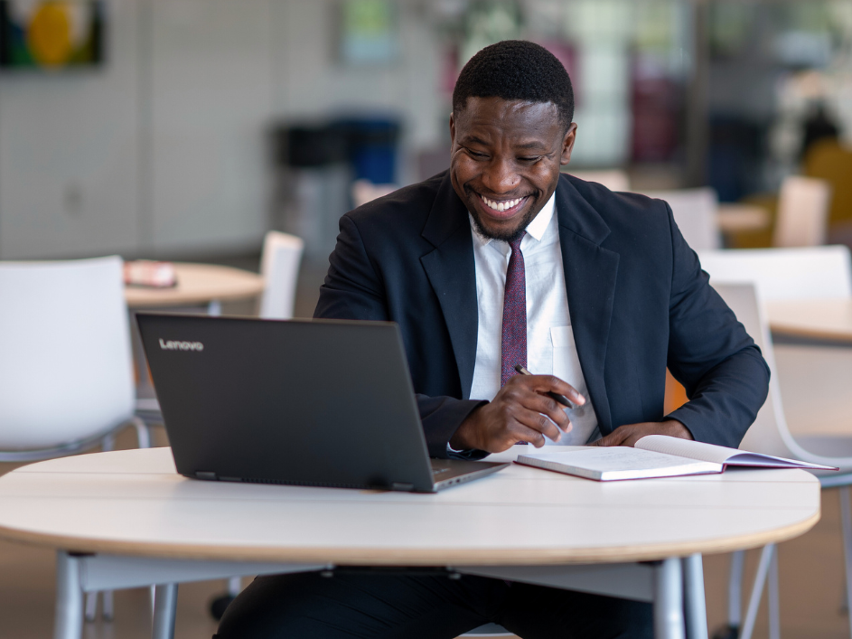 A man in a suit smiles while looking at a laptop and writing in a notebook at a table in a bright, open space.