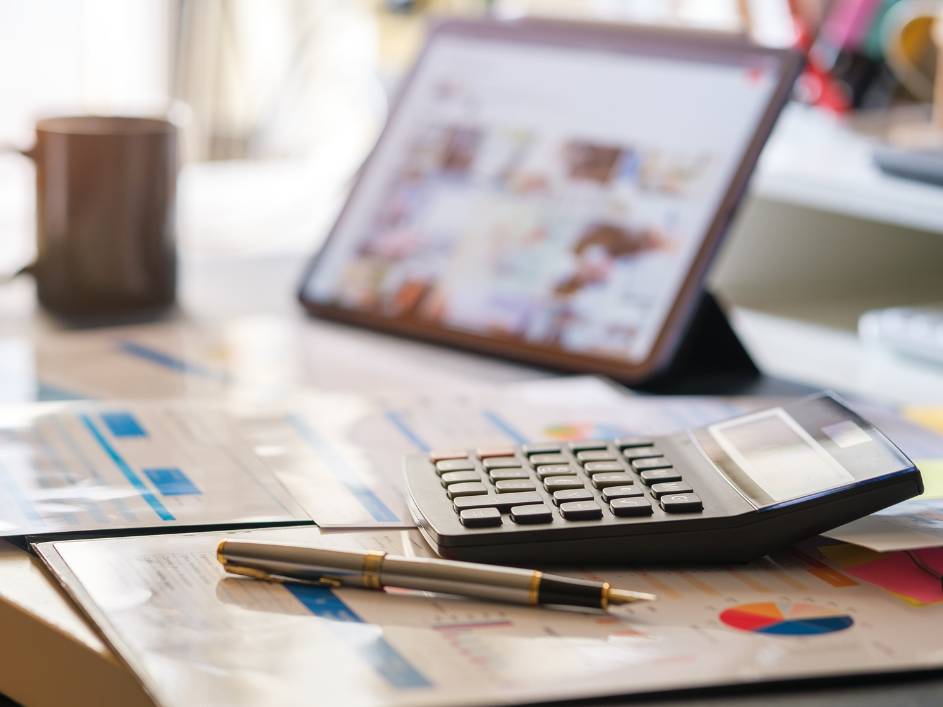 A mug, calculator, pen, and papers with papers on a desk, with a blurry tablet in the background.