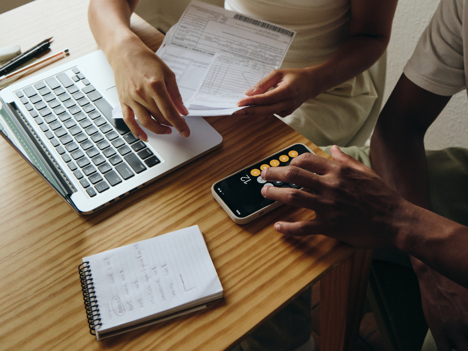 A top-down view of two people at a desk, one holding papers, the other using a calculator on a phone, with a laptop and notebook nearby.