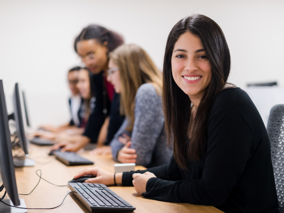 A female student in a computer lab smiles at the camera, with other students on computers blurred in the background.