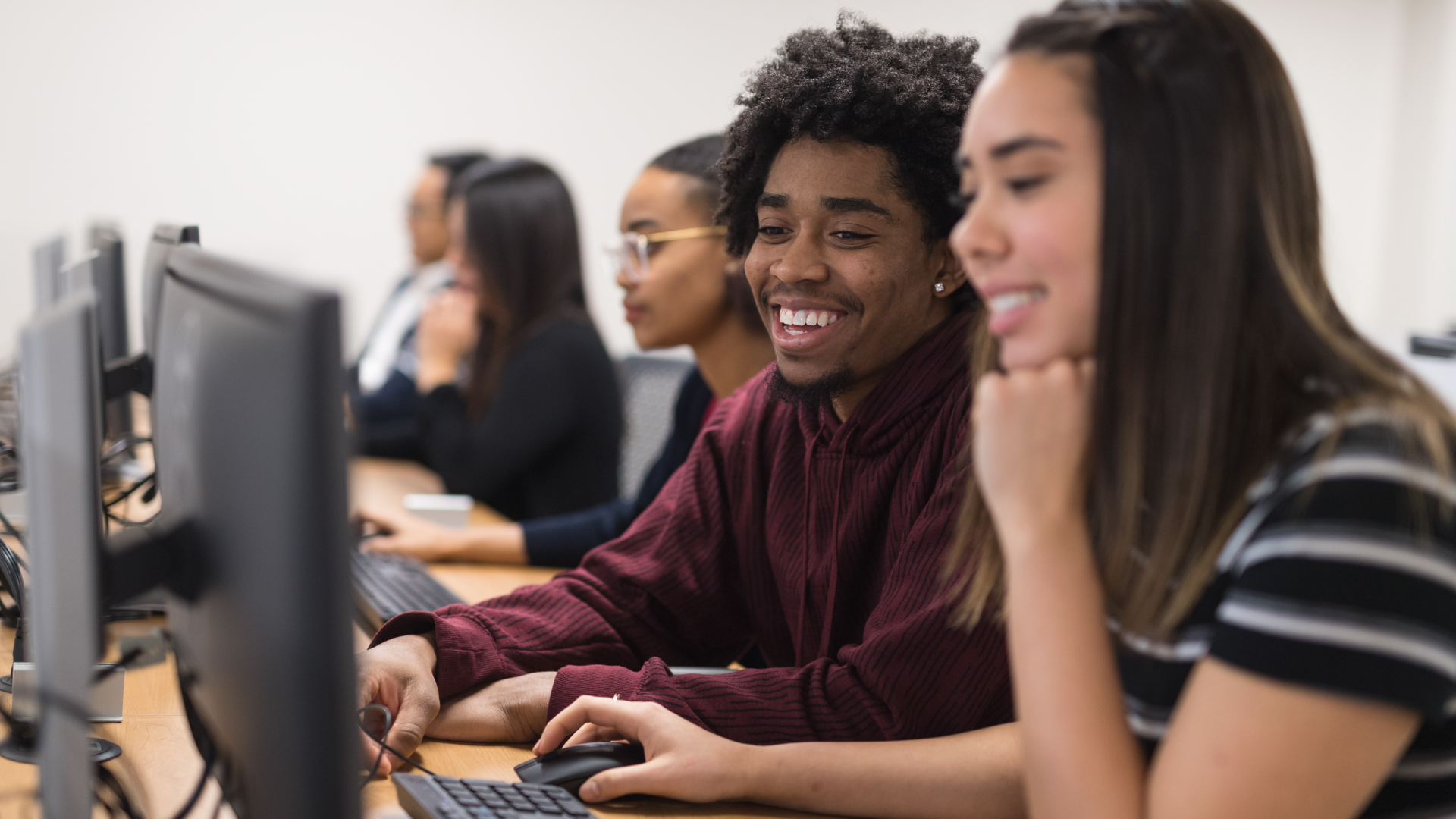 A young man with curly hair smiles while working on a computer next to a young woman, with other students in the background.