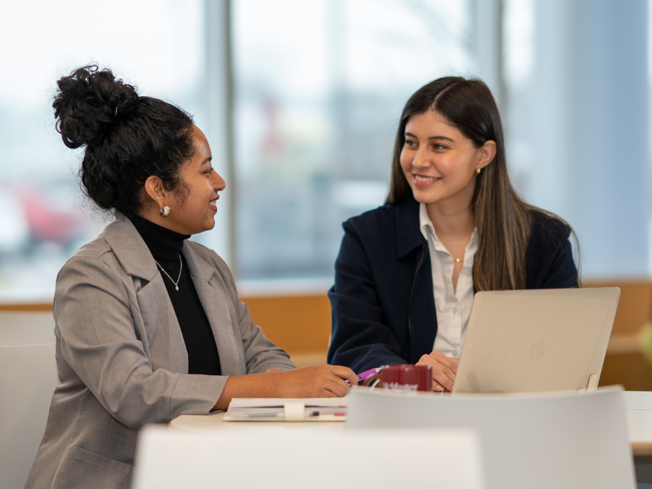 Two smiling female students, one with dark curly hair and one with long straight hair, converse across a table with a laptop.