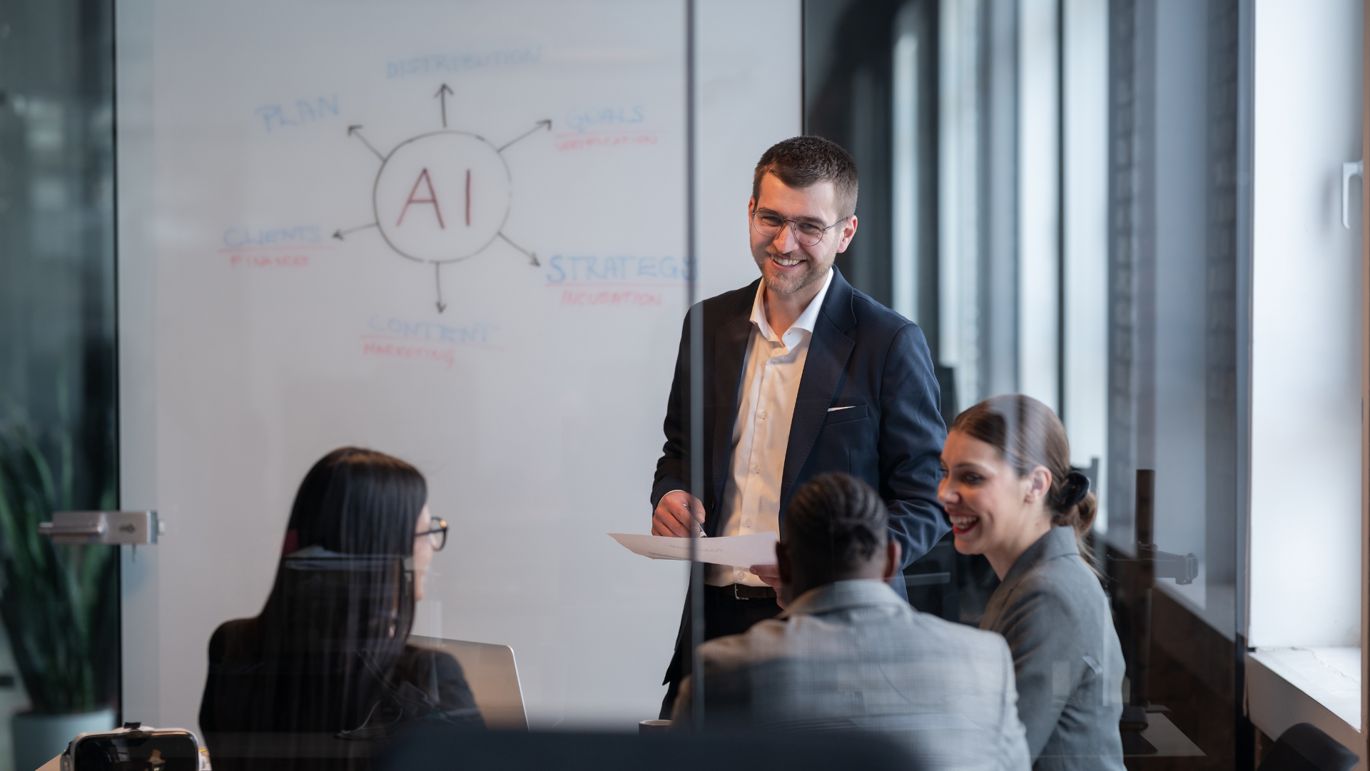 A male presenter in a suit smiles and holds papers while standing in front of a whiteboard with an "AI" diagram.